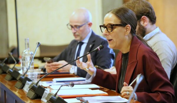 La Sindaca Metropolitana Sara Funaro relaziona al microfono; seduta al tavolo ingombro di appunti; sullo sfondo il capo di gabinetto