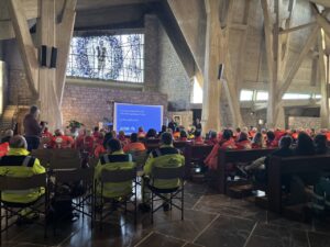 interno della chiesa sull'autostrada, pavimento marmo marrone e alti pilastri di cemento; sullo sfondo vetrata; su panche e sedie i volontari con giubbotti gialli e arancioni ripresi di schiena