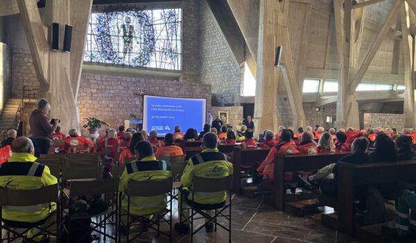 interno della chiesa sull'autostrada, pavimento marmo marrone e alti pilastri di cemento; sullo sfondo vetrata; su panche e sedie i volontari con giubbotti gialli e arancioni ripresi di schiena