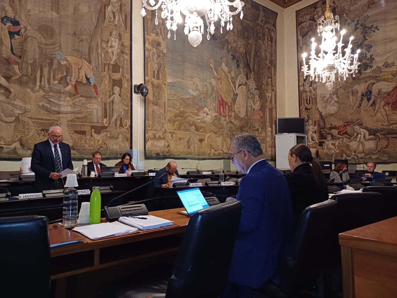 veduta d'angolo della sala 4 stagioni durante il consiglio; 3 arazzi alle pareti e 2 lampadari di cristallo; agli scranni i consiglieri seduti, in piedi interviene il consigliere Armentano