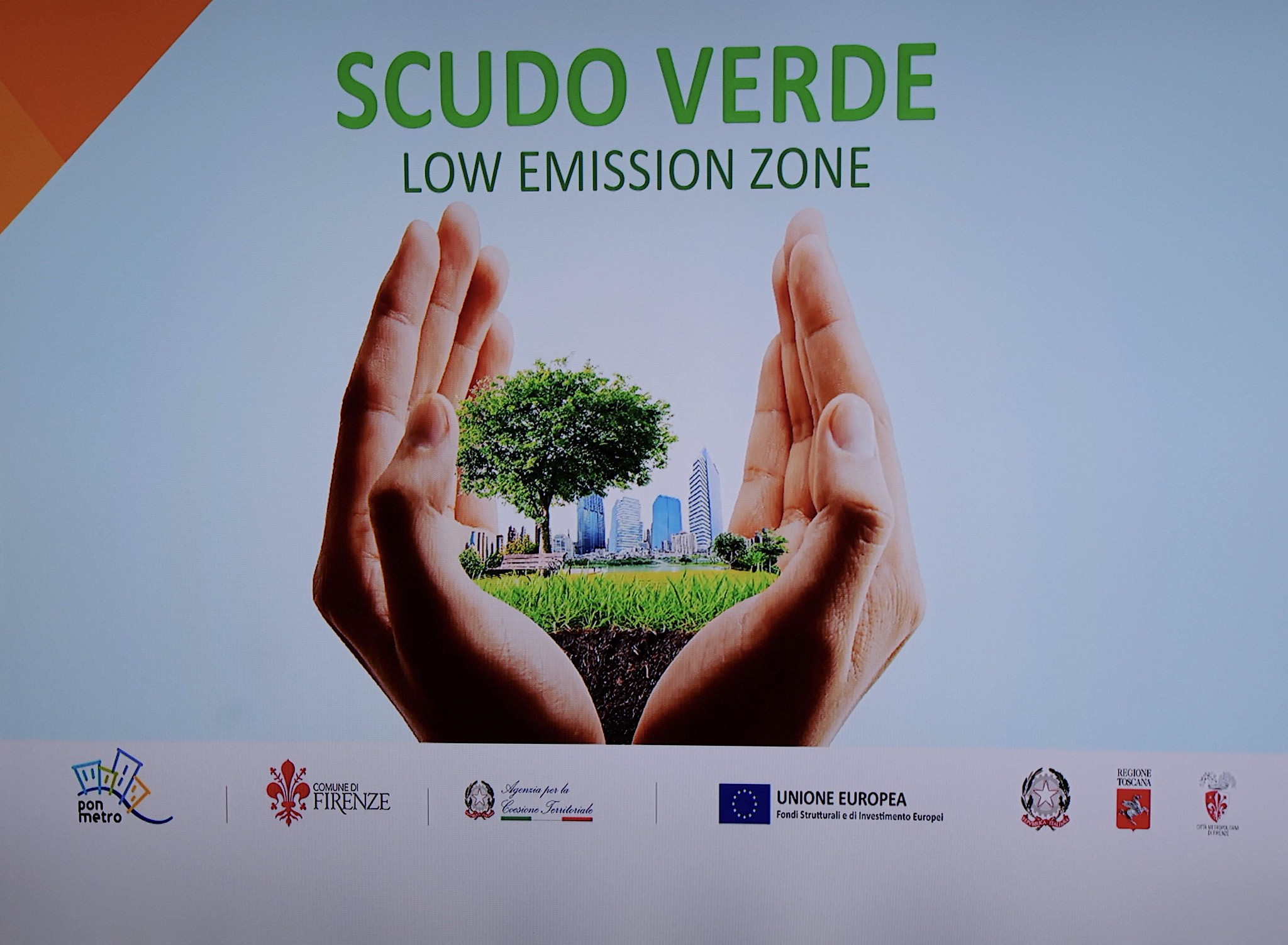 scudo verde low emission zone, fra due mani a conca è contenuto un parco in primo piano e abitazioni sullo sfondo