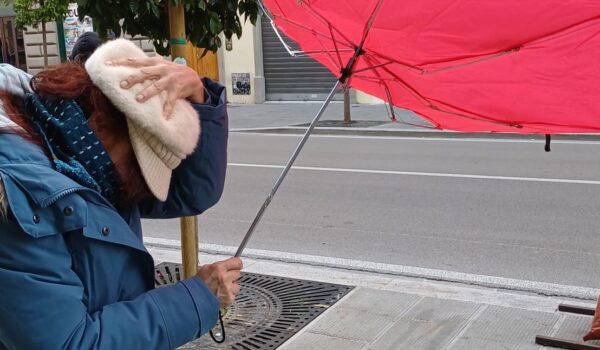 scorcio di una via cittadina, in primo piano una donna che con una mano tiene il cappello sulla testa, e con l'altra regge l'ombrello che si è rigirato per il forte vento