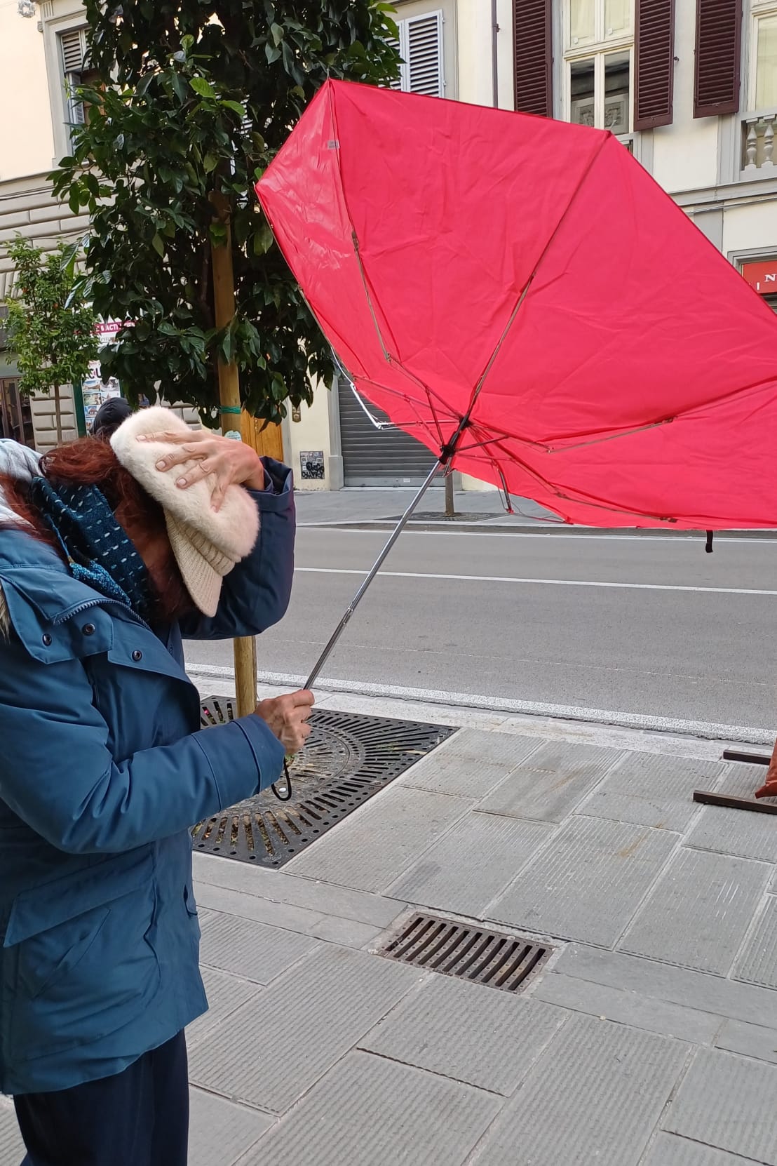 scorcio di una via cittadina, in primo piano una donna che con una mano tiene il cappello sulla testa, e con l'altra regge l'ombrello che si è rigirato per il forte vento